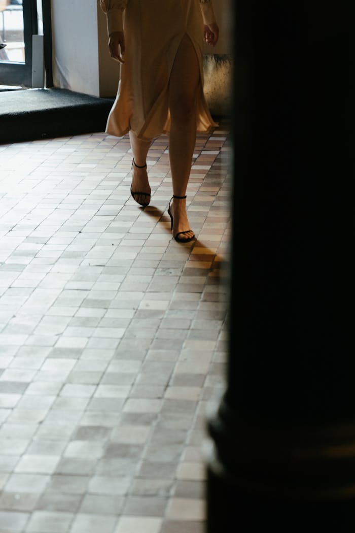 Stylish woman in a flowing dress and high heels walking indoors on a tiled floor.